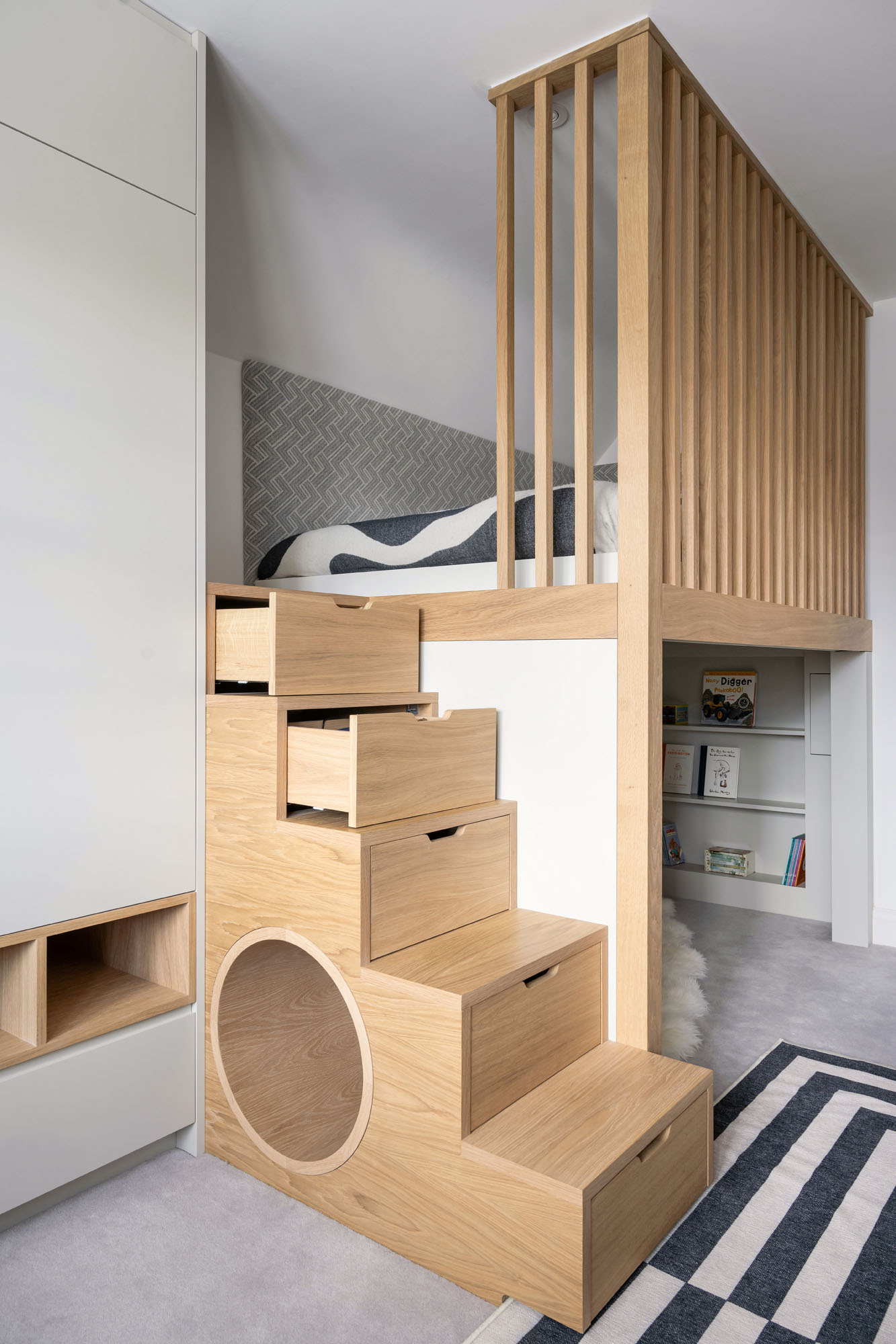 A bespoke boys’ bedroom by Funky Little Darlings in Amersham, showcasing a raised loft bed with oak steps featuring built-in storage drawers. A fun play tunnel adds an adventurous touch leading to a reading nook blending playful design with practical functionality.
