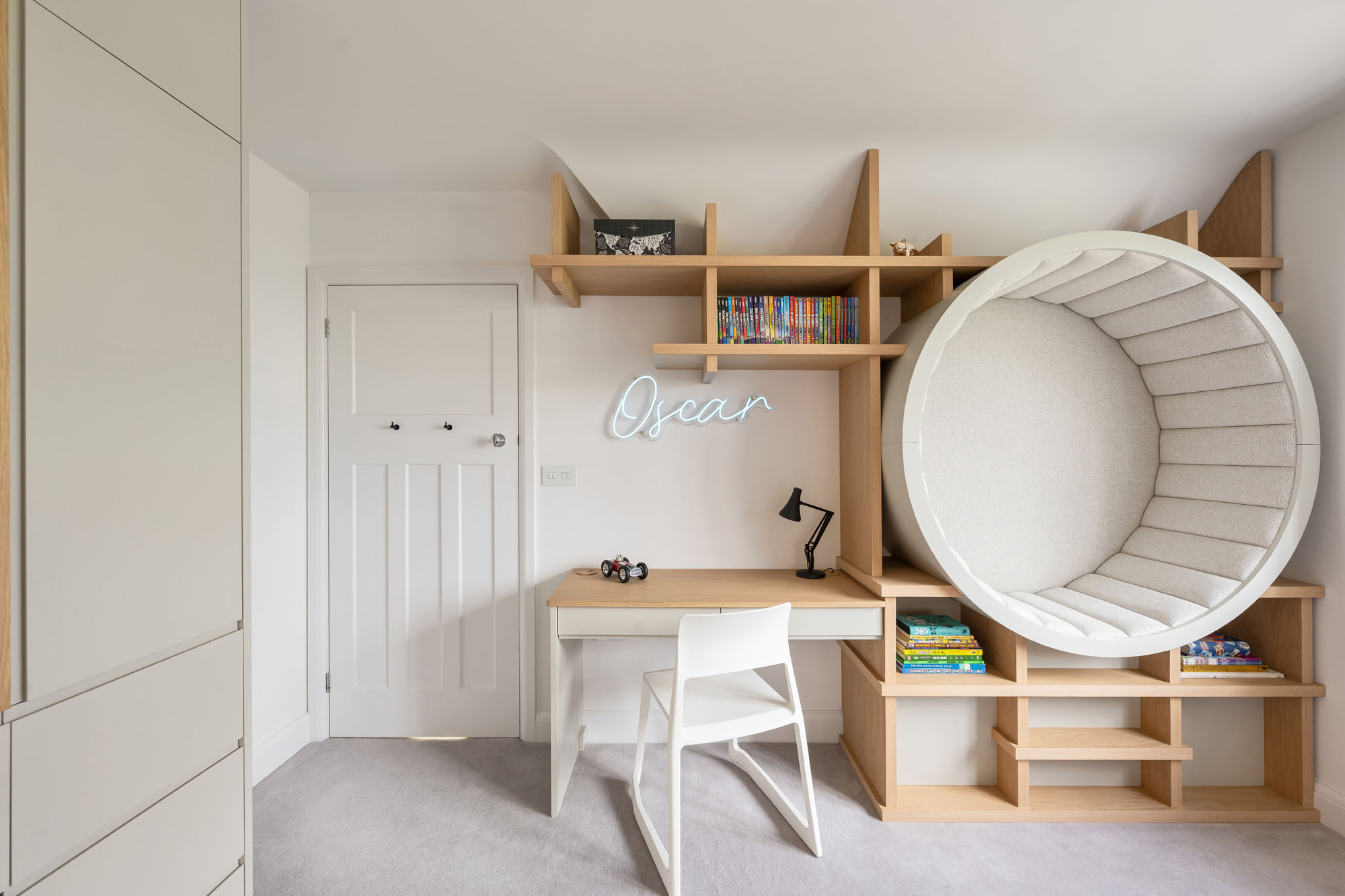 Modern and imaginative boy's bedroom in Amersham, designed by Funky Little Darlings, showcasing a stylish Vitra desk chair, bespoke neon name sign, open wall-mounted storage, and a cosy upholstered circular seat for relaxing in.