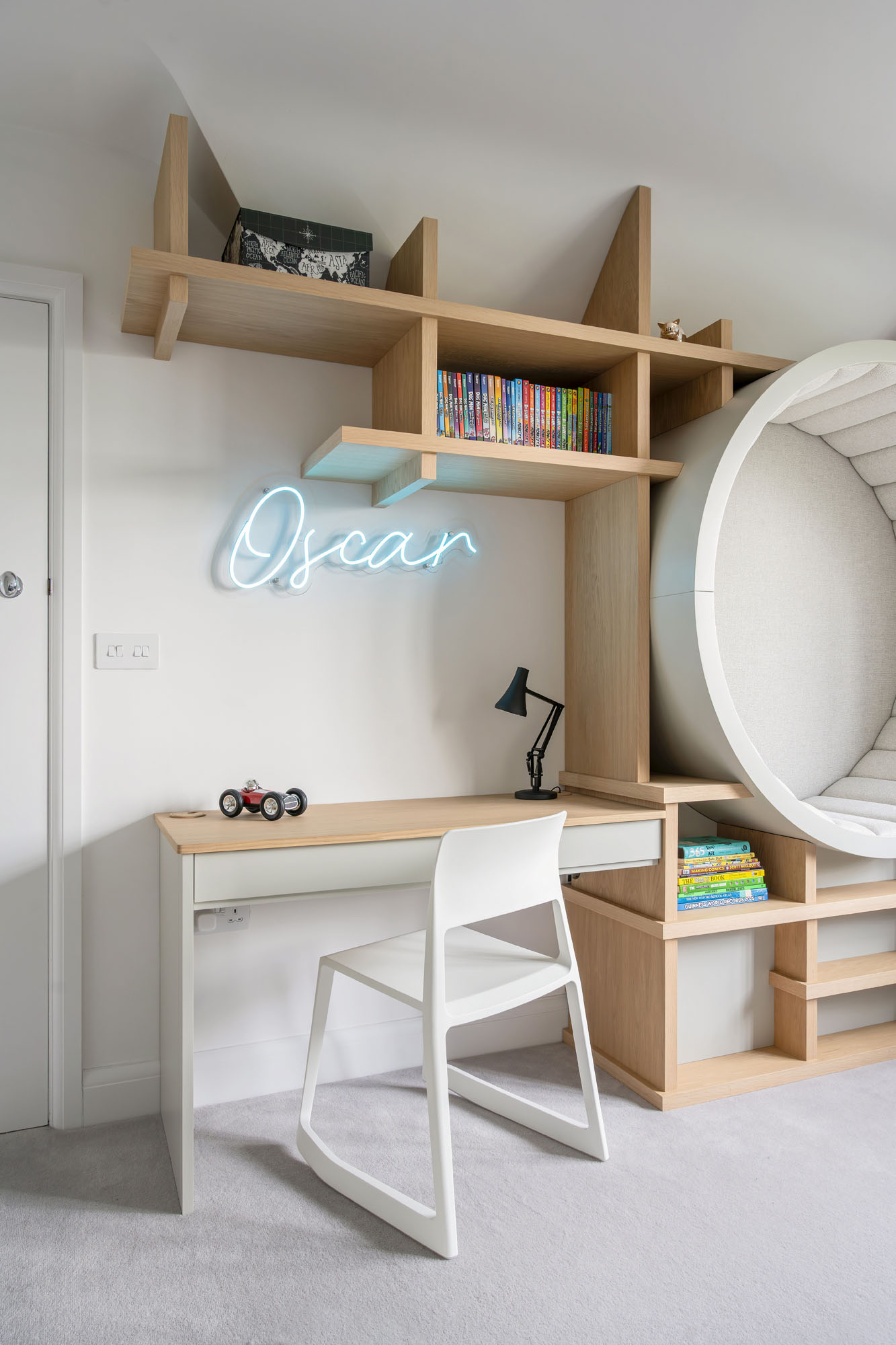 Modern and minimalist boy’s bedroom designed by Funky Little Darlings in Amersham, featuring a sleek study desk with a white contemporary chair, Vitra-style design, custom wooden shelving with open storage, a personalised neon 'Oscar' sign, and a unique bespoke circular upholstered seat built into the joinery, combining functionality, creativity, and personalised design for a stylish children’s interior.