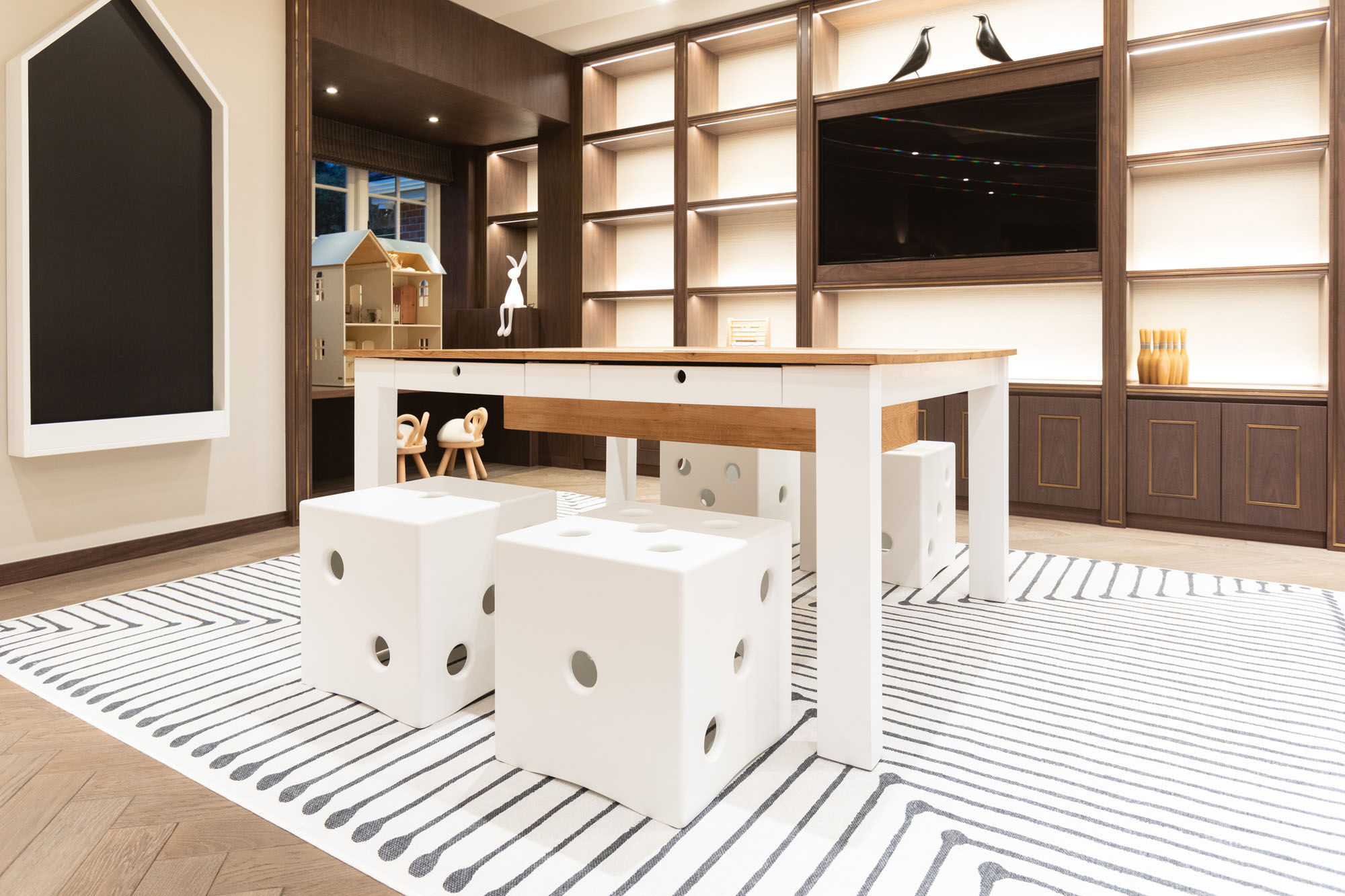 Contemporary children’s playroom in Cobham, Surrey, designed by Funky Little Darlings, featuring a house-shaped chalkboard, craft table with white dice shaped stools and integrated TV display with stylish storage solutions, perfect for creative and functional family living.