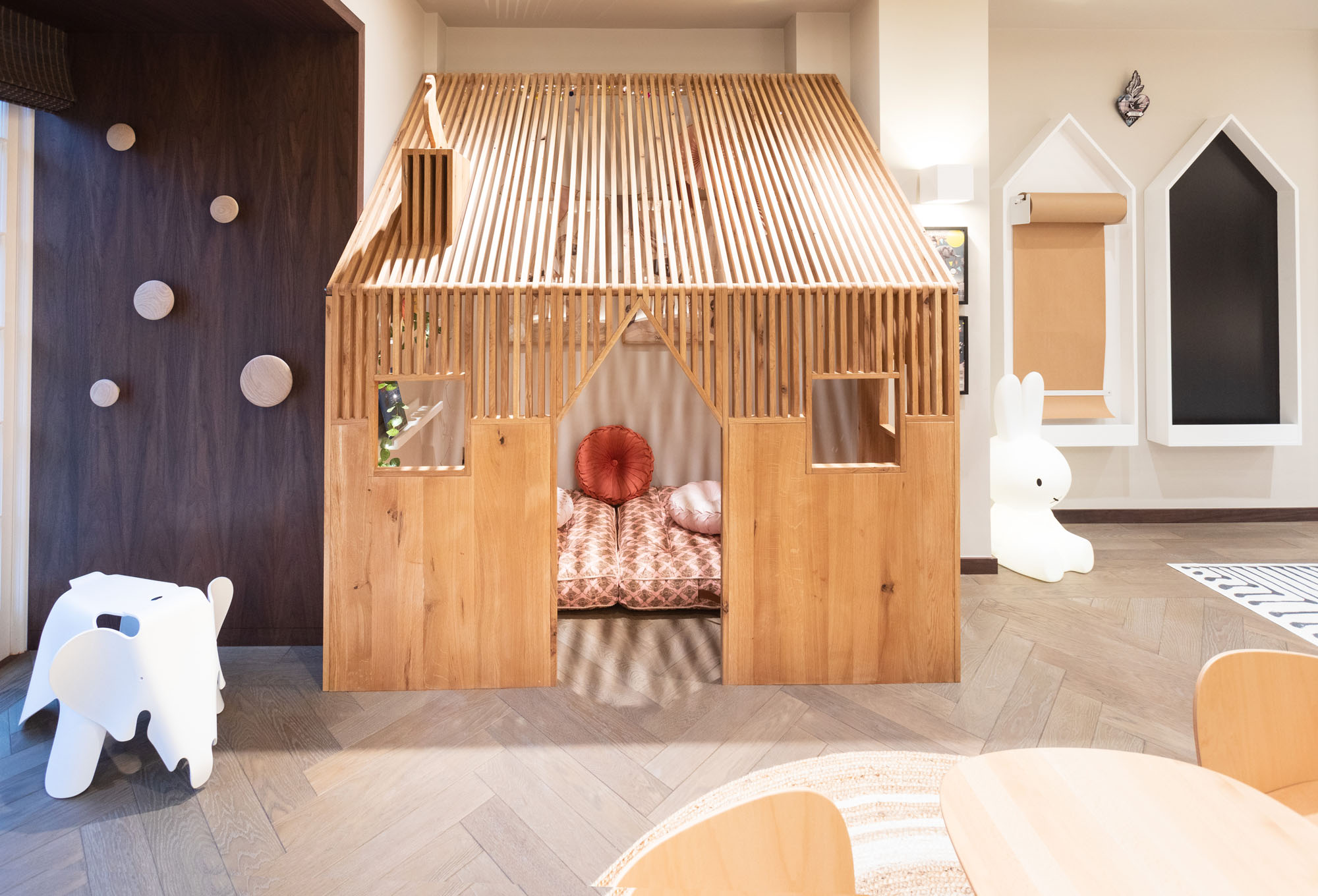 Bespoke children’s playroom in Cobham designed by Funky Little Darlings, featuring a whimsical indoor playhouse complete with pigeon on the chimney, iconic Eames elephant stool, Miffy floor lamp and wall-mounted house shaped paper roll holder and chalkboard. A playful space fostering imagination and creative play.