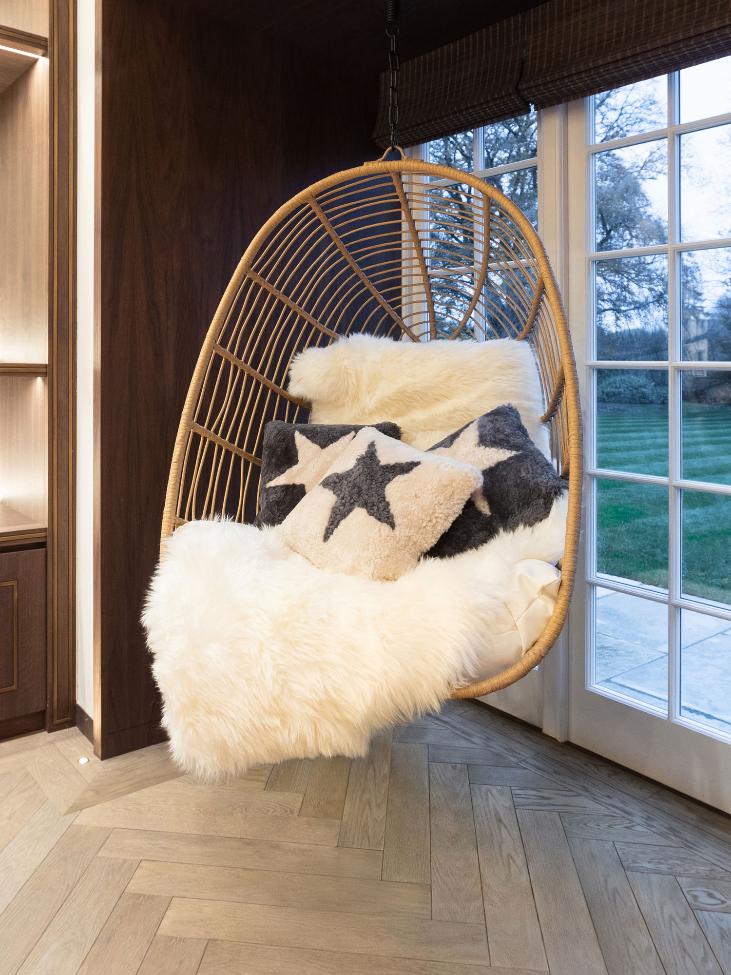 Modern children's playroom in Cobham by Funky Little Darlings featuring a cosy hanging swing chair piled with sheepskins and cushions, bespoke display shelves and stylish designer furniture, blending creative play with contemporary kids' interiors.