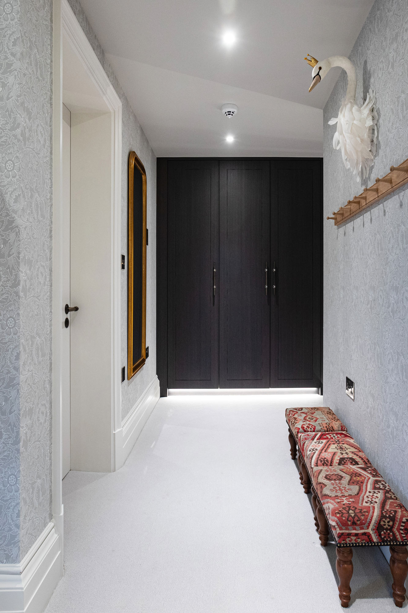 Elegant shared triple children's bedroom in Cobham by Funky Little Darlings, featuring bespoke built-in wardrobe cabinetry, classic William Morris floral wallpaper, row of stools outside the bathroom for the children in this thoughtfully designed layout for stylish and functional sibling living.