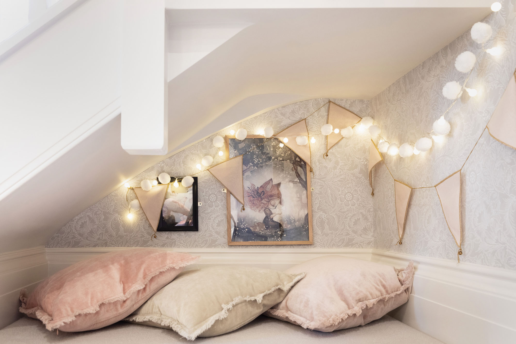 Whimsical shared triple kids' bedroom in Cobham designed by Funky Little Darlings, featuring a cosy secret den under the stairs with velvet cushions, twinkling fairy lights, bunting with bells and classic William Morris floral wallpaper for a magical and stylish space.