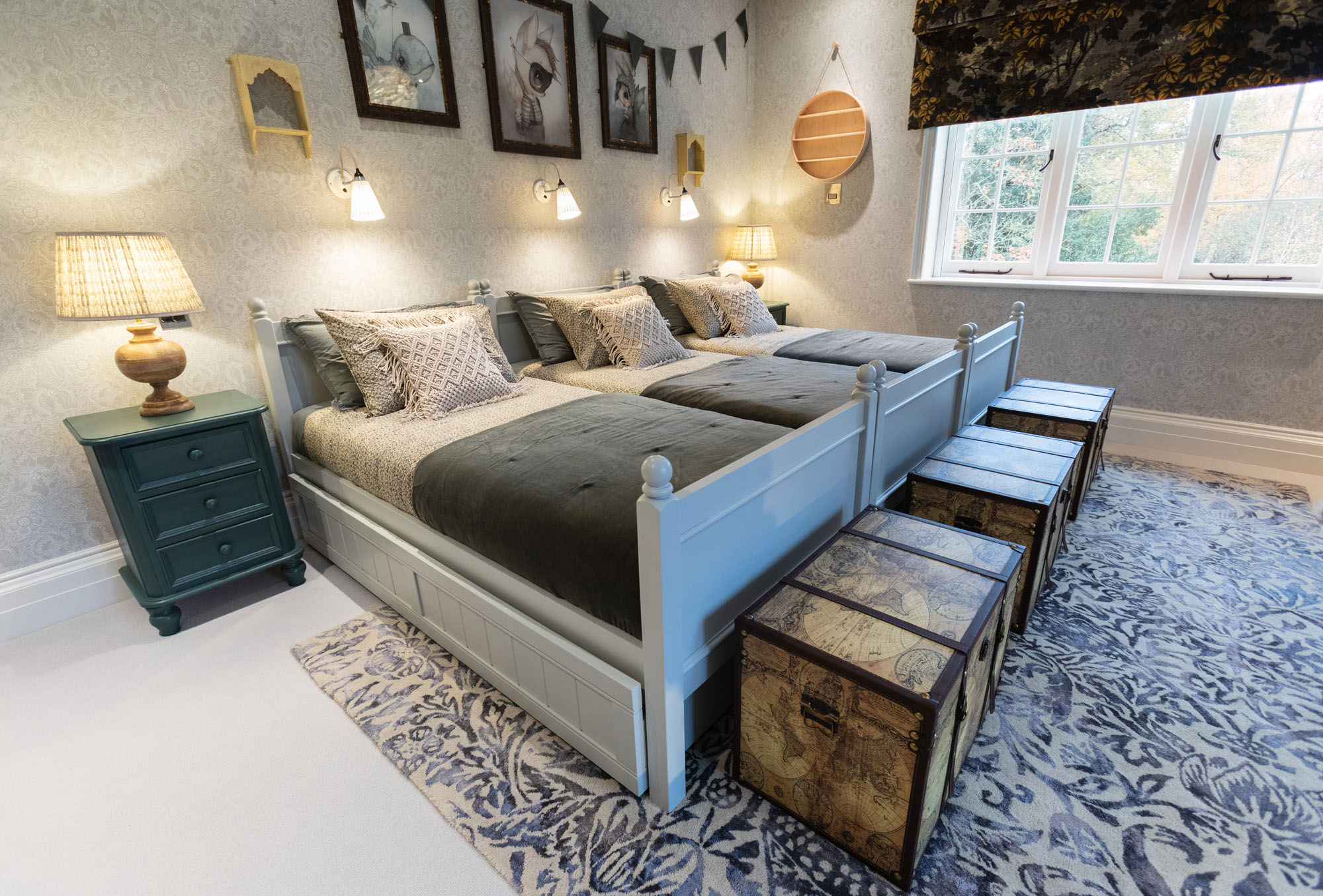 Charming shared triple bedroom in Cobham designed by Funky Little Darlings, featuring three single beds with vintage-style storage trunks, vibrant Pooky table lamps, classic William Morris wallpaper, and a patterned rug that adds a nostalgic, heritage-inspired vibe to this stylish children’s space.