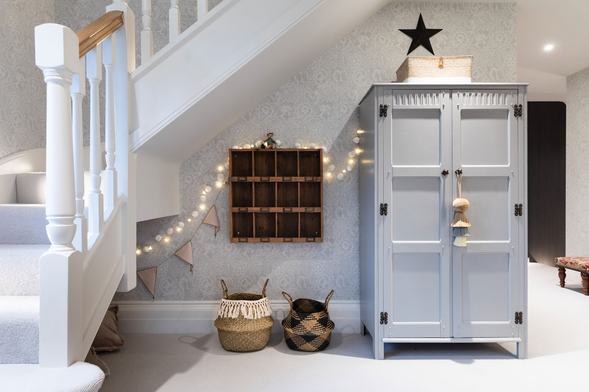 Funky Little Darlings Cobham shared triple bedroom showcasing a hand-painted vintage wooden wardrobe with intricate detailing, a rustic wooden shelf, woven belly baskets for organised storage, soft glowing fairy lights draped across the room and elegant William Morris floral wallpaper creating a cosy and whimsical children’s bedroom atmosphere.