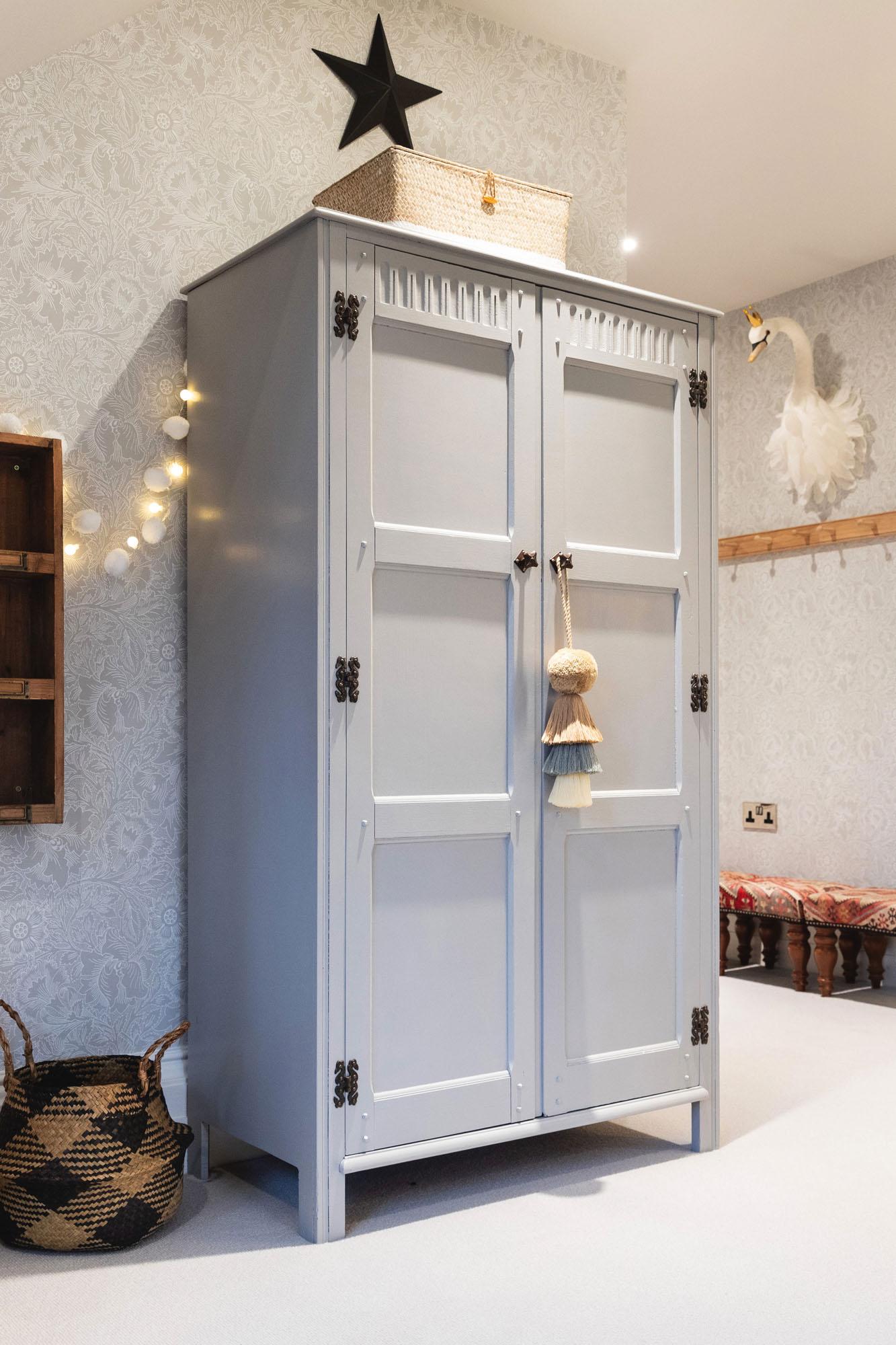 Funky Little Darlings Cobham shared triple bedroom showcasing a hand-painted vintage wooden wardrobe with intricate detailing, a rustic wooden shelf, woven belly baskets for organised storage, soft glowing fairy lights draped across the room and elegant William Morris floral wallpaper creating a cosy and whimsical children’s bedroom atmosphere.