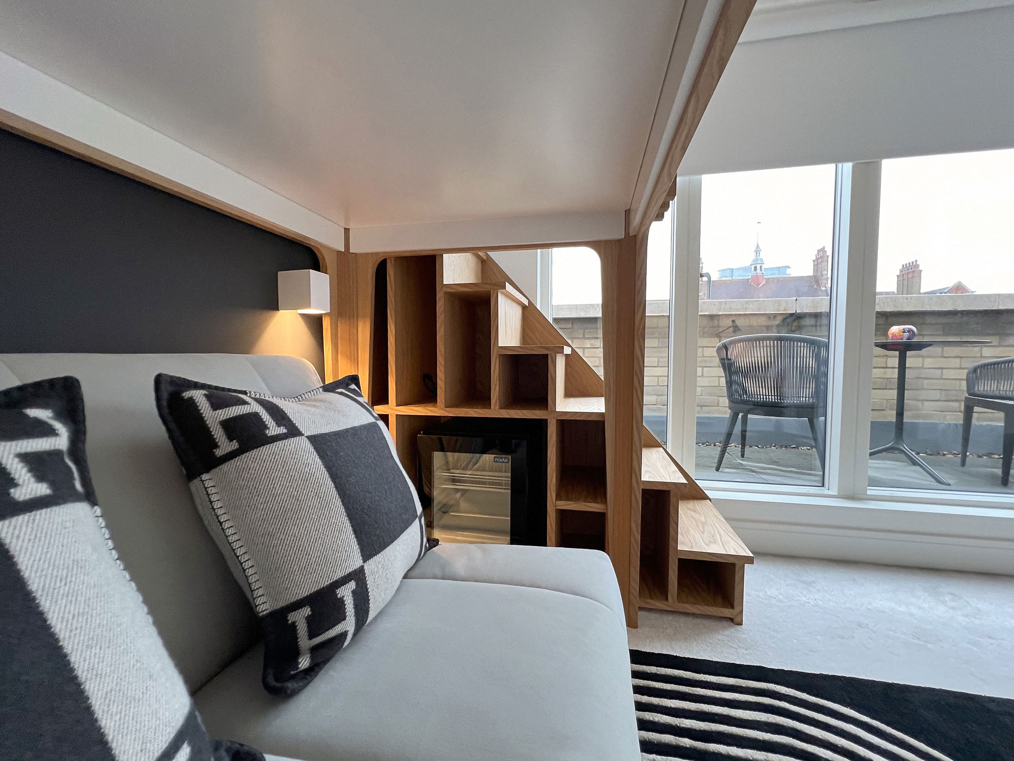 View of Fulham boys’ bedroom designed by Funky Little Darlings, featuring a white bespoke loft bed with oak details and steps, sofa bed with Hermès cushions, wall lights, compact fridge integrated into built-in storage, black graphic rug and access to a balcony.