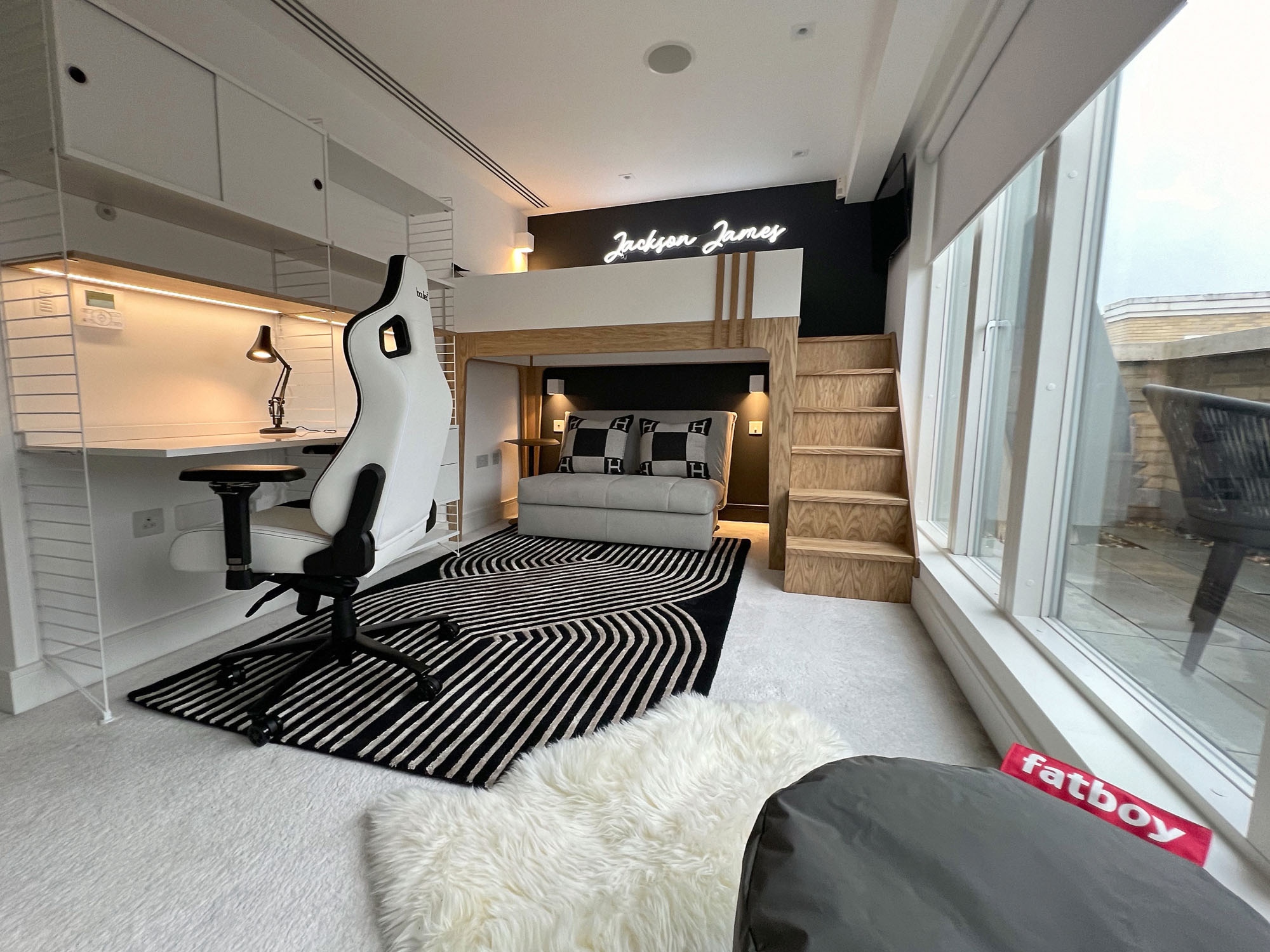 Stylish Fulham boys' bedroom by Funky Little Darlings featuring a custom white bespoke loft bed with oak details, vibrant neon sign, and cosy sofa bed accented with Hermes cushions. This playful yet practical space includes modern wall lights, a mini fridge, String shelving system with desk and storage, a sleek gaming chair, and a bold black graphic rug with sheepskin and grey Fatboy pouffes, perfect for a design-forward, teen-friendly interior.