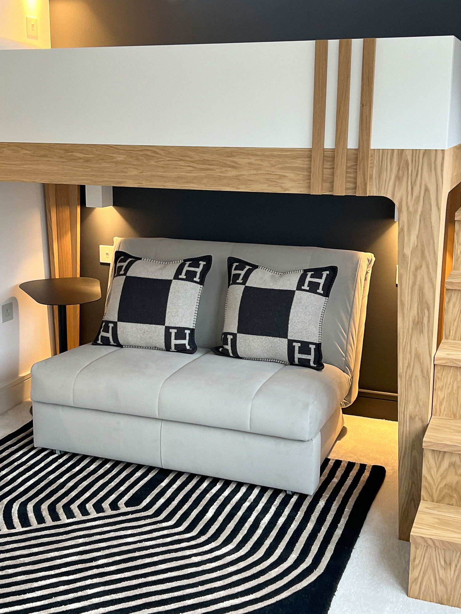 Contemporary boy's bedroom in Fulham designed by Funky Little Darlings, featuring a white bespoke loft bed with oak details, grey sofa bed with Hermès cushions, Neptune Charcoal wall paint, integrated wall lights, side table and a bold black graphic rug.