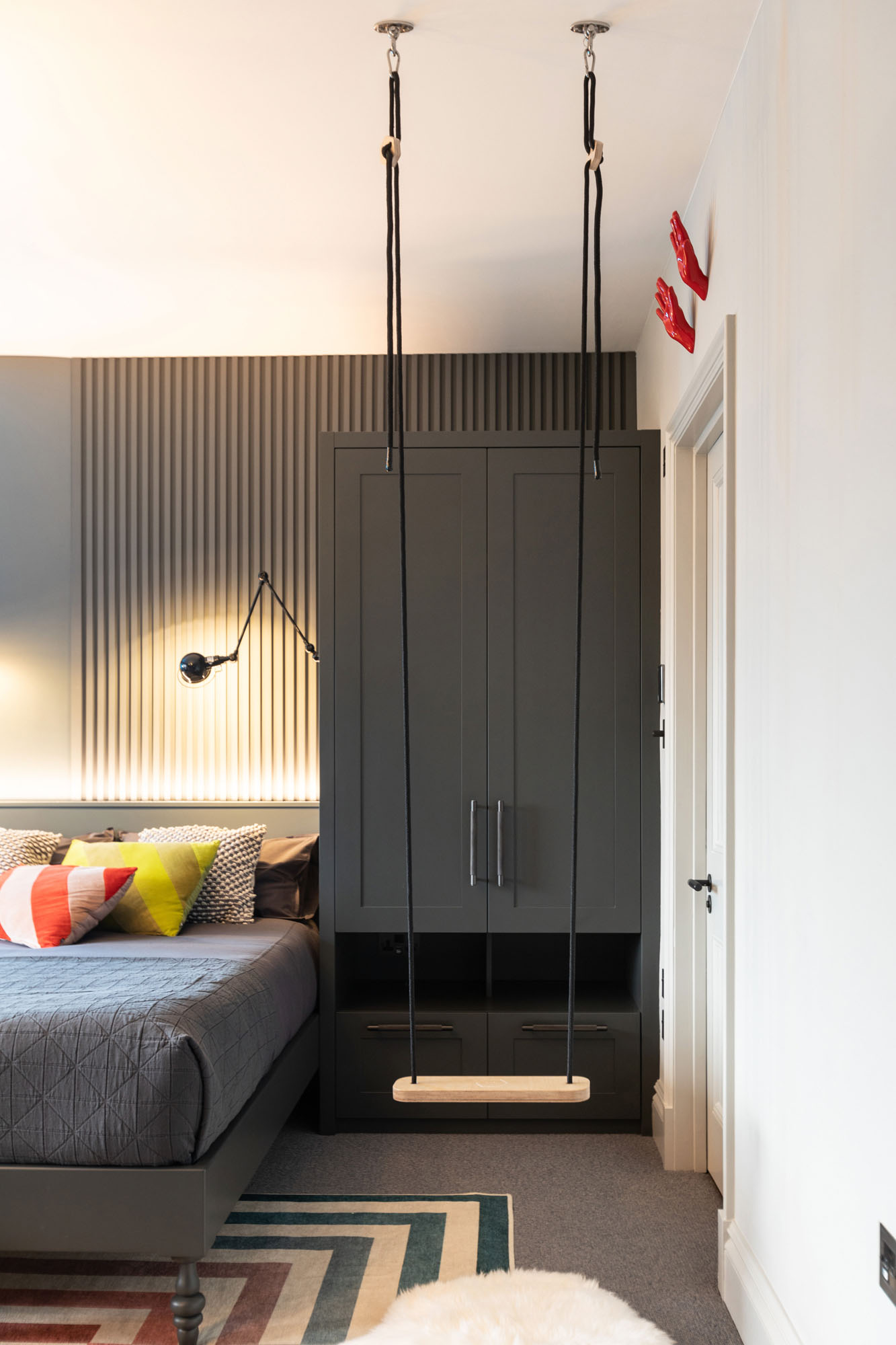 Contemporary Hampstead boy’s bedroom designed by Funky Little Darlings featuring custom built-in wardrobes with ample storage, a bespoke bed crafted for comfort and style, sleek modern slatted wood wall panelling adding texture, a playful hanging swing chair suspended from the ceiling, and industrial-style Jielde wall lights providing functional and stylish illumination. This design perfectly blends practical storage solutions with bold, playful elements to create a unique, design-led children’s bedroom interior.